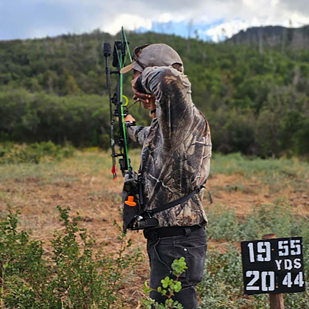 Person in camouflage gear with a bow and arrow in an outdoor setting, with a target distance sign in the foreground.